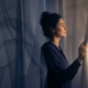 A thoughtful woman parting translucent curtains and looking toward warm light