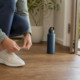 A person ties walking shoes beside an open doorway in the morning, representing a kind and confident first step toward healthy habits.