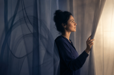 A thoughtful woman parting translucent curtains and looking toward warm light